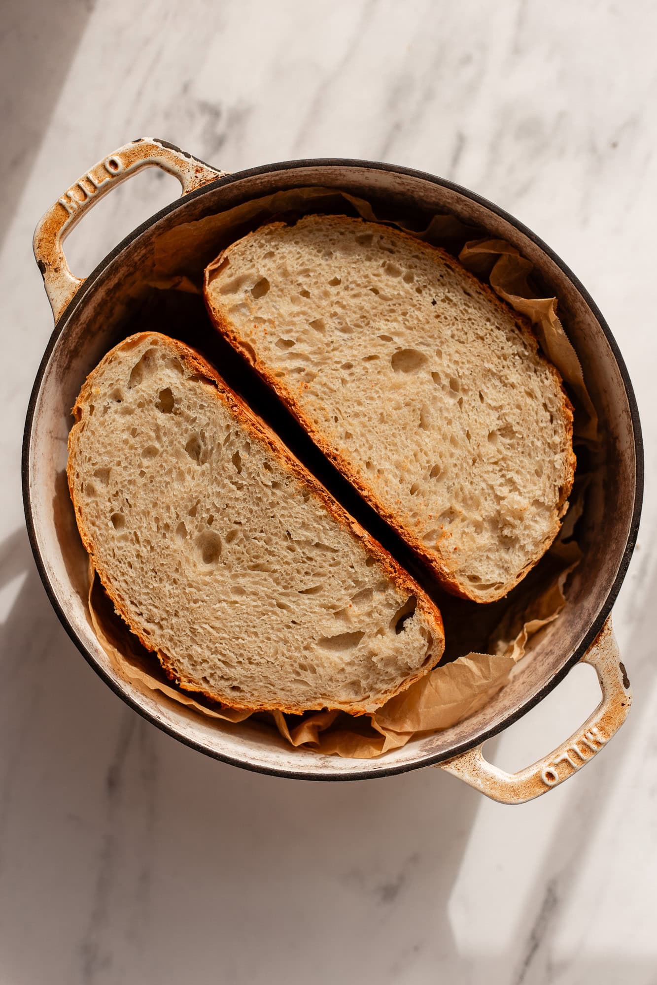 A sourdough boule sliced in half, showing its airy crumb structure, resting in a Dutch oven lined with parchment paper.