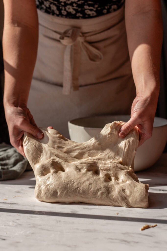 Hands stretching sourdough dough to check elasticity on a clean, unfloured surface, emphasizing hydration and technique.