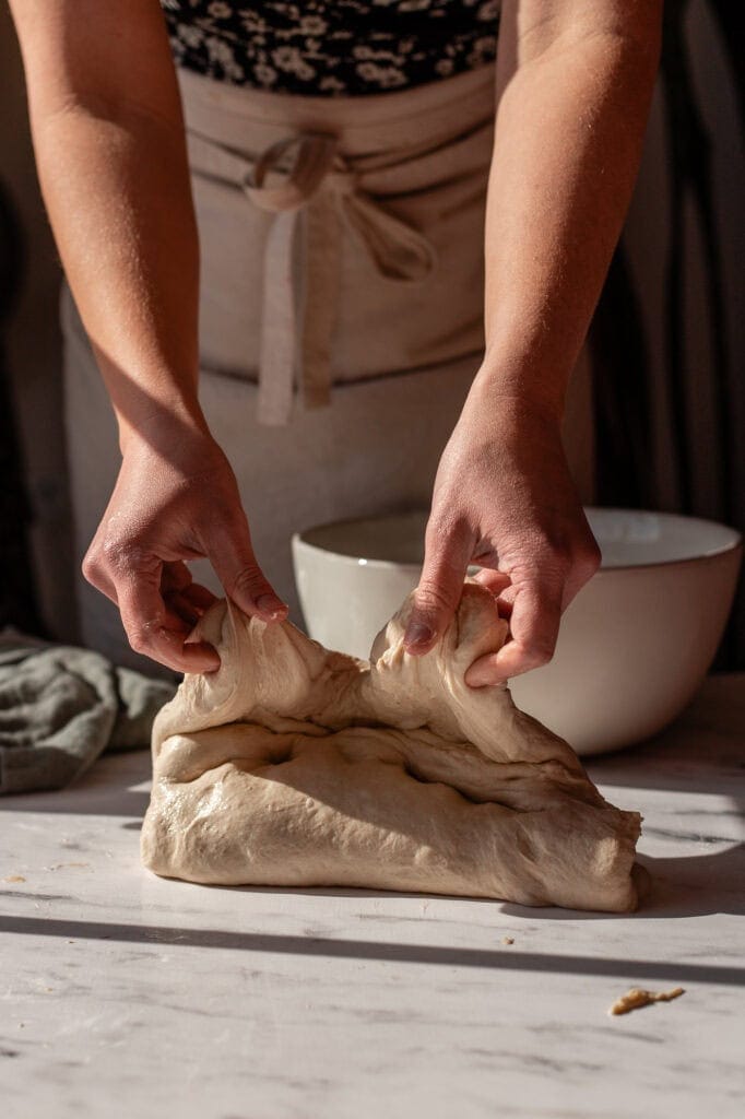 Hands gently pulling sourdough dough upward to start the folding process, preparing for structure building.