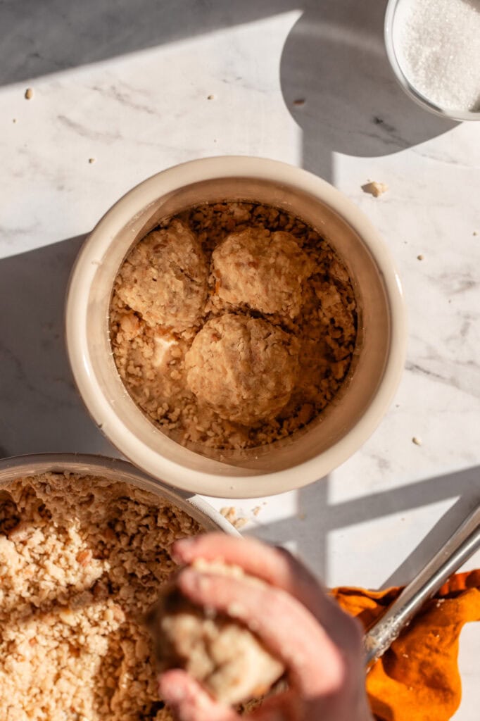 Top-down view of a ceramic crock with miso balls placed inside, surrounded by loose miso mixture. A hand prepares to throw another miso ball into the crock to ensure a snug fit and eliminate air pockets.