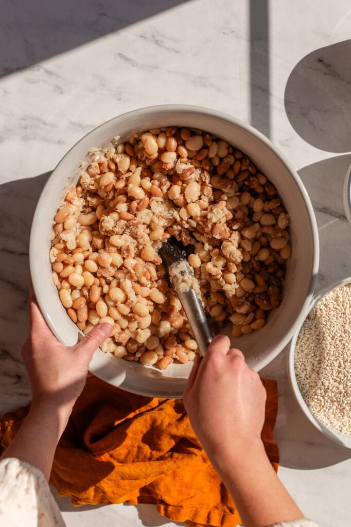 Hands using a masher to mash a mixture of cooked white beans in a large ceramic bowl, preparing them for soy-free miso paste. 