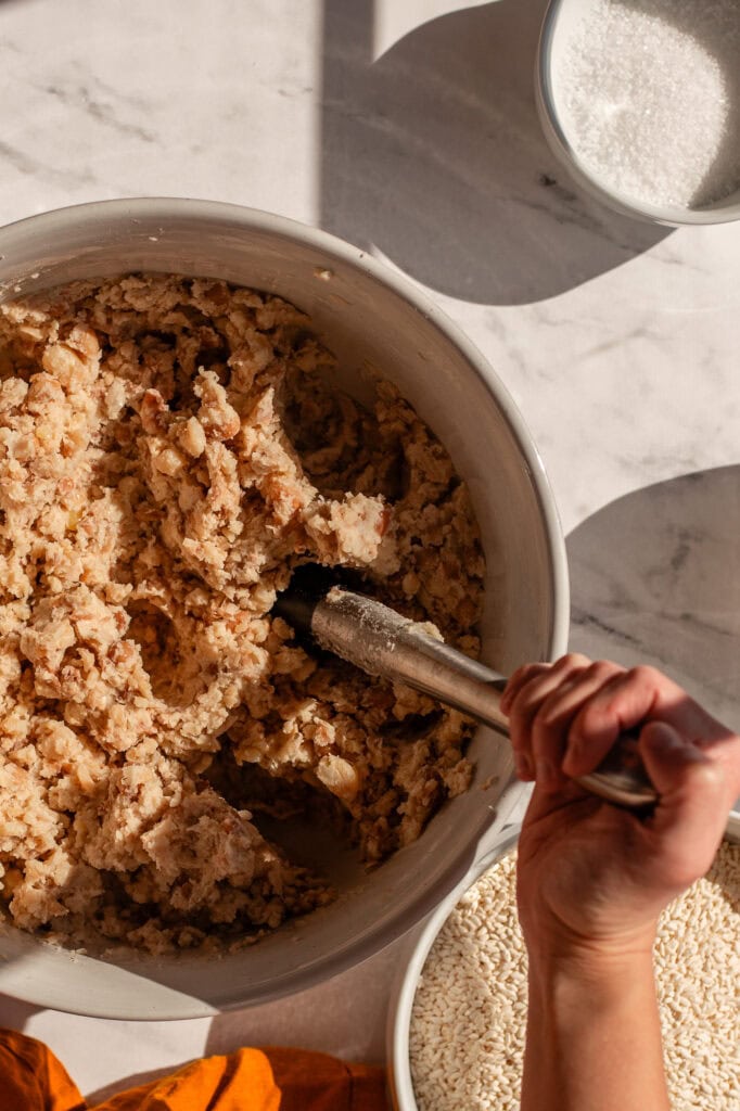 Close-up of mashed beans in a ceramic bowl, showing a textured consistency as they are further broken down with tongs. Koji rice and salt are visible in bowls nearby.