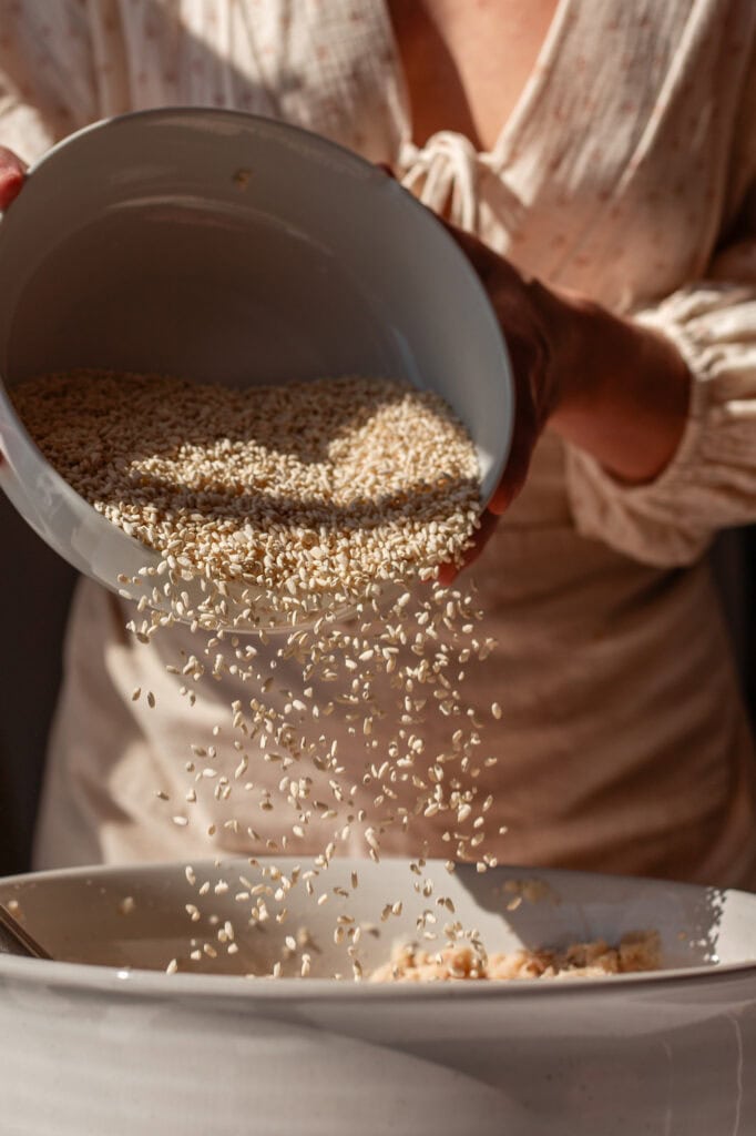 Hands pouring koji rice from a white bowl into a larger mixing bowl filled with mashed beans, capturing the motion of the grains cascading down.