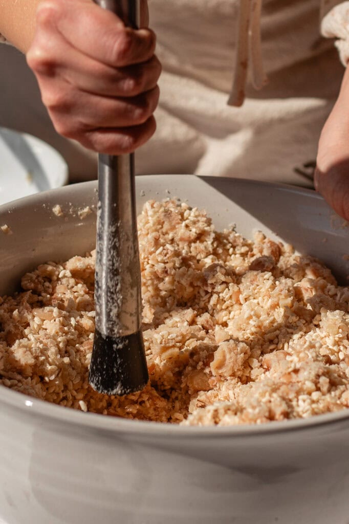 Hands using a masher to mix koji rice into mashed beans in a large ceramic bowl, preparing the base for soy-free miso paste. 