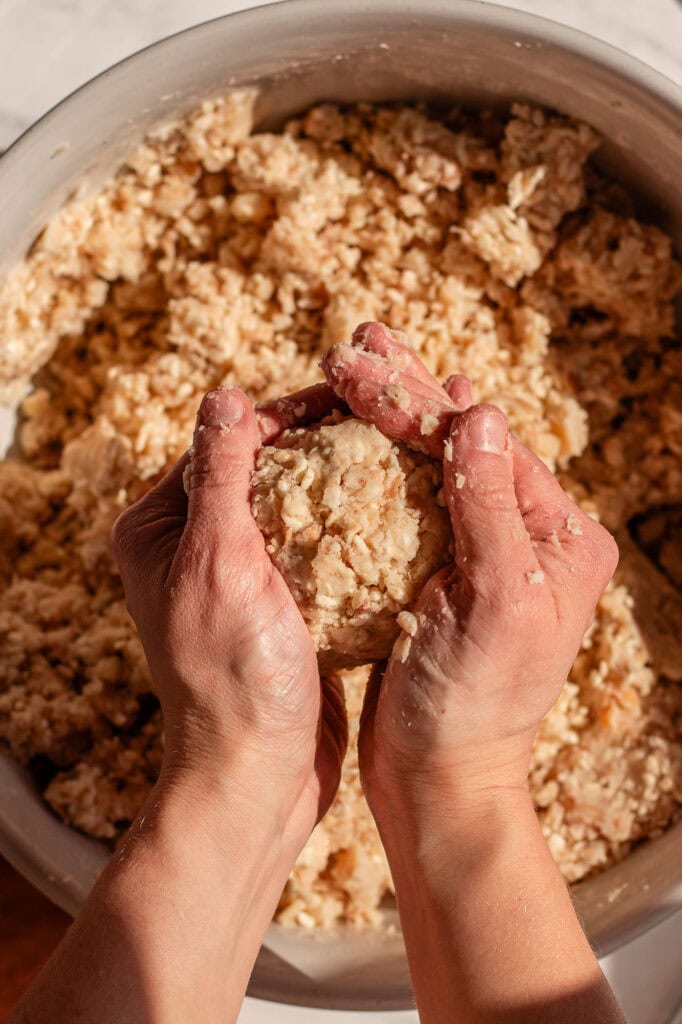 Hands pressing together a portion of miso mixture, forming a compact miso ball. The textured mixture of mashed beans, koji rice, and salt is visible in a large bowl in the background.