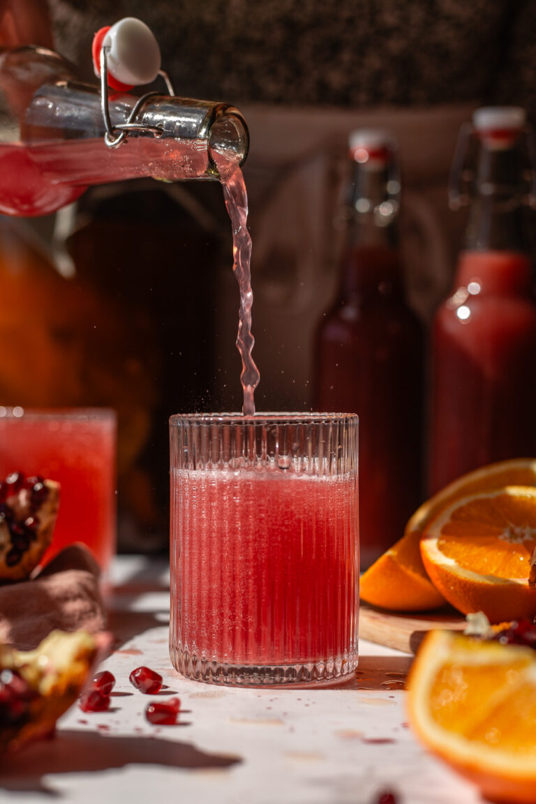 A finished bottle of pomegranate kombucha pouring into a small glass.