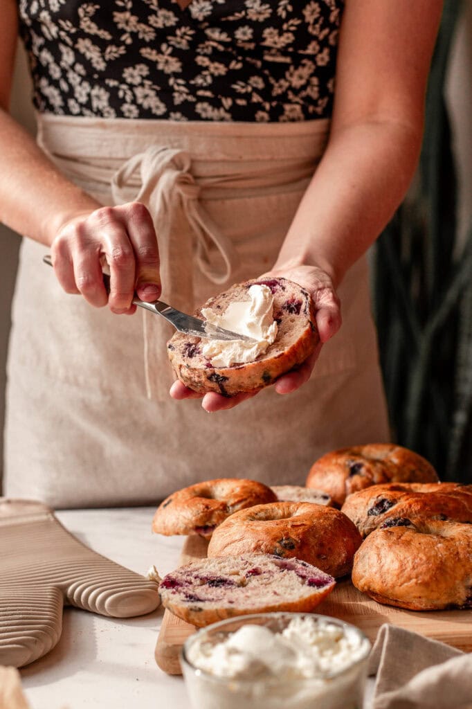 Hands holding a sourdough blueberry bagel and using a knife to spread cream cheese across one half.