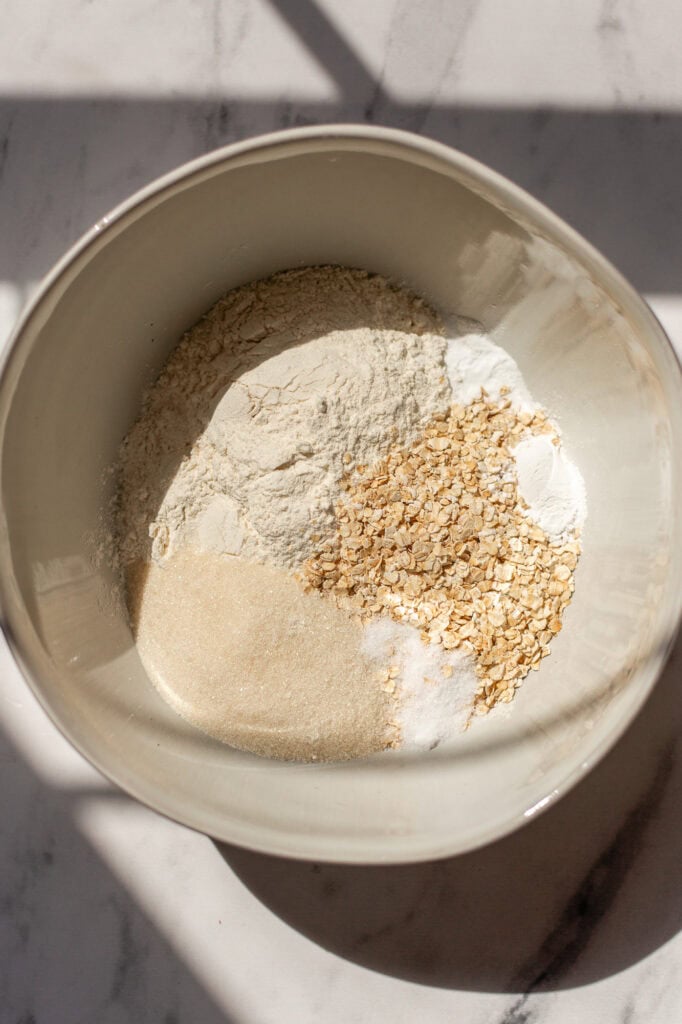 A ceramic mixing bowl filled with dry ingredients for oatmeal strawberry muffins, including flour, rolled oats, sugar, baking powder, and salt.