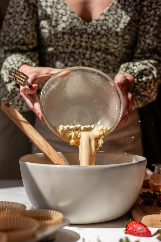 A person wearing a floral blouse pours a bowl of wet ingredients—melted butter, sourdough starter, lemon juice, whole milk, and eggs—into a larger mixing bowl.