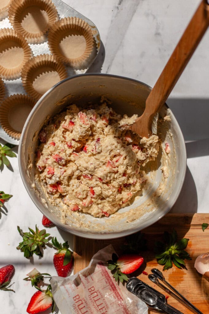 A mixing bowl filled with thick, textured oatmeal strawberry muffin batter, speckled with vibrant red strawberry pieces. 