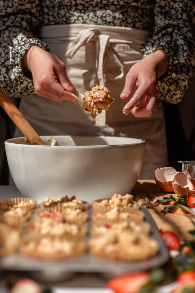 A person spoons oatmeal strawberry muffin batter into parchment-lined muffin cups. 