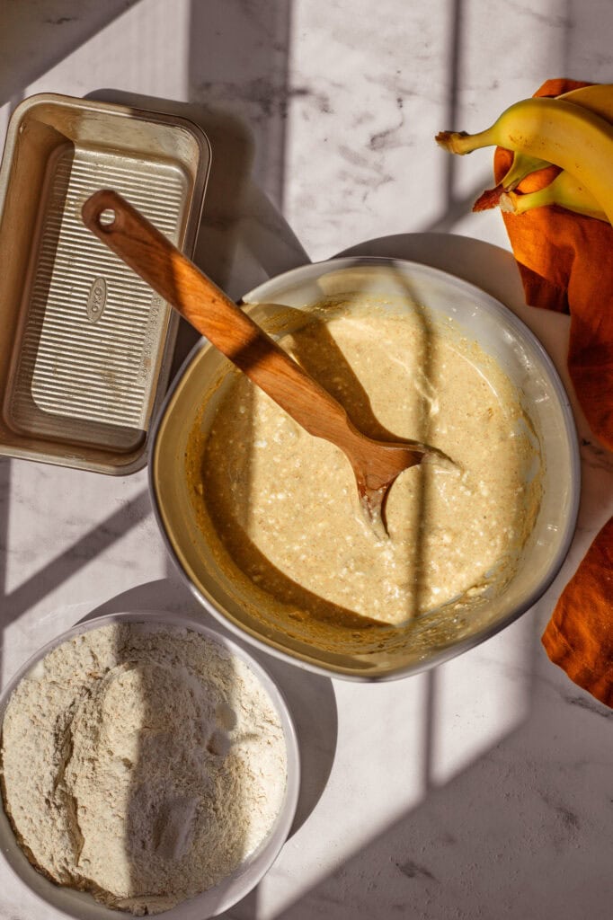 Overhead view of a mixing bowl with wet ingredients for cottage cheese banana bread, next to a separate bowl of dry ingredients including flour, protein powder, and spices.