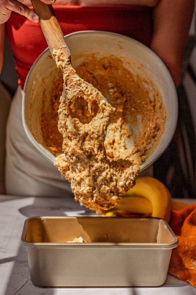 A wooden whisk pushes cottage cheese banana bread batter into a butter coated loaf pan.