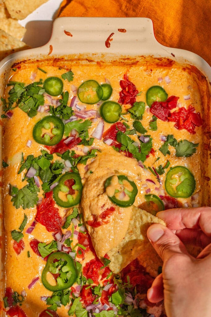 A hand dipping a tortilla chip into a hot, creamy cottage cheese queso dip topped with jalapeños, roasted red peppers, red onion, and fresh cilantro.
