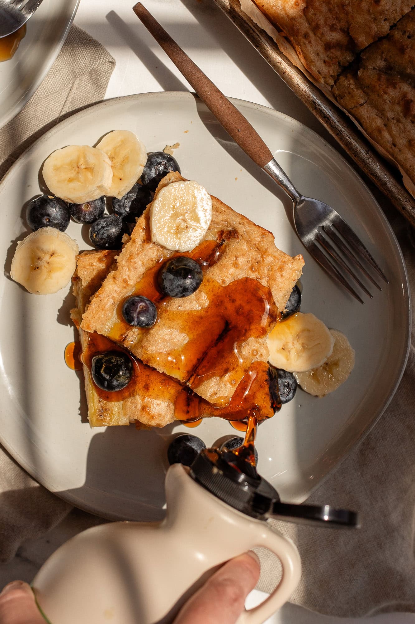 Stack of sourdough cottage cheese pancakes topped with banana slices, blueberries, and maple syrup being poured from a bottle.