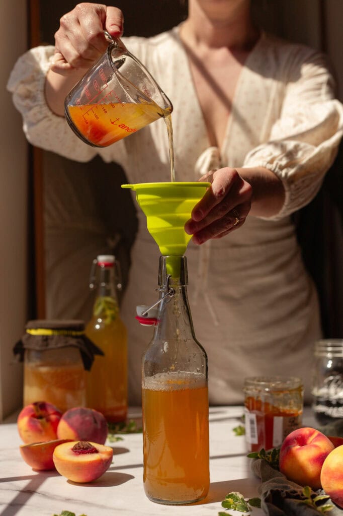 Hands pouring a peach preserve mixture into a bottle of kombucha primary ferment.