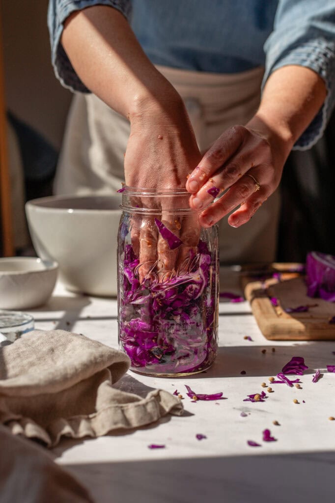 Hands pressing vibrant red cabbage and blueberries into a glass jar for fermentation, with scattered coriander seeds on a white marble counter.