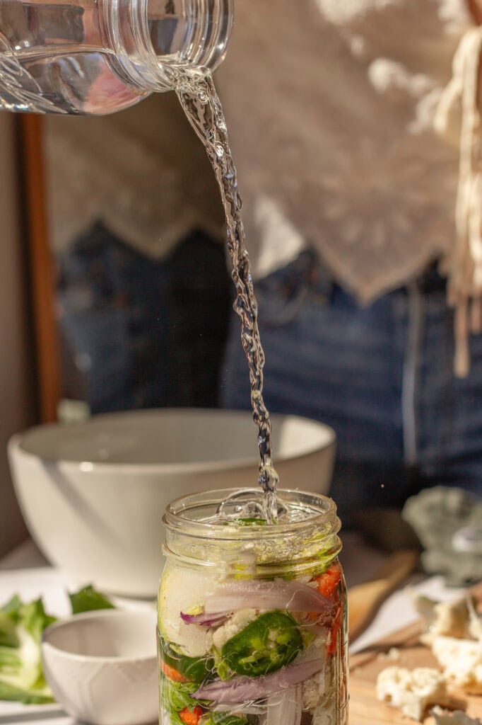 Using a temper to press down vegetables in a jar A jar filled with vegetables and water being poured into it.
