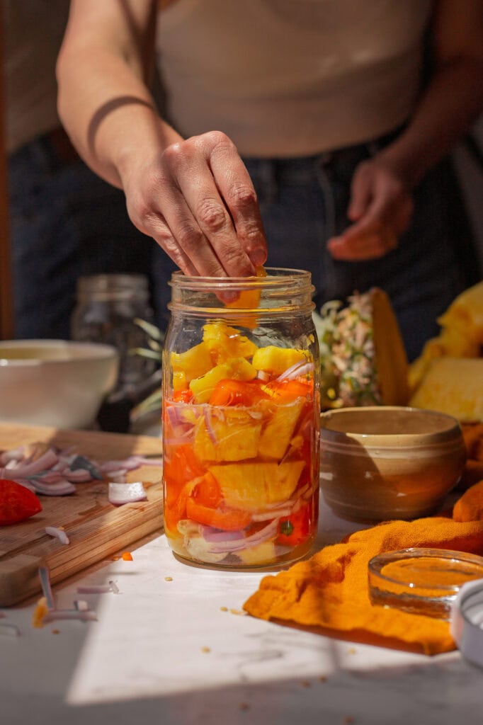 Hands placing chopped pineapple and peppers into a jar. 