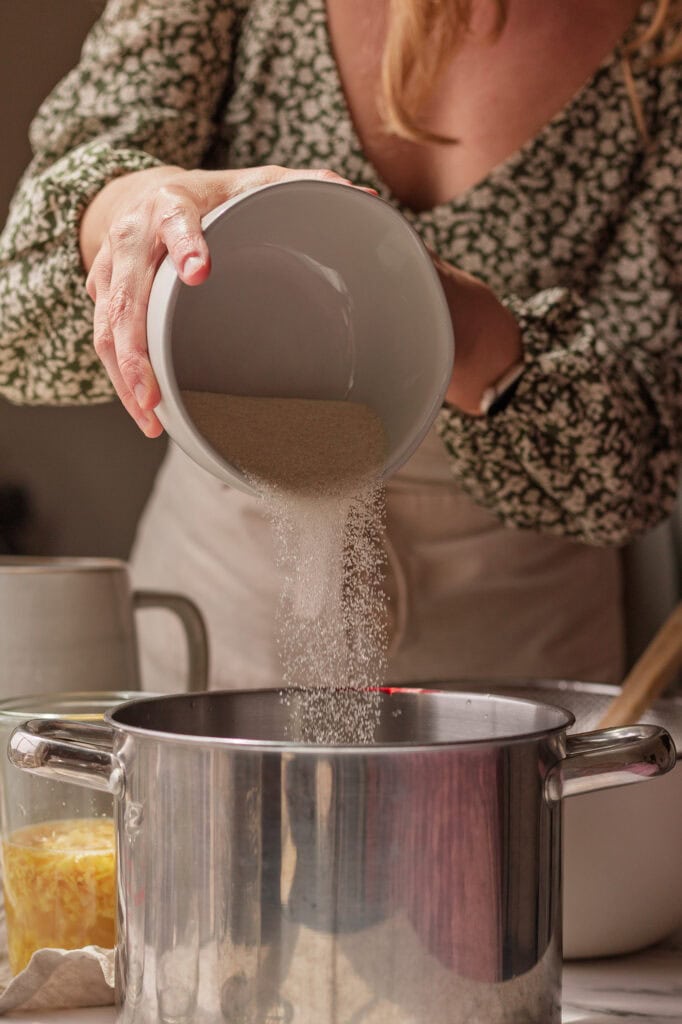 Pouring Sugar Into Ginger Bug Drink Mix Hands pouring sugar into a stainless steel pot, adding to the lemon juice.