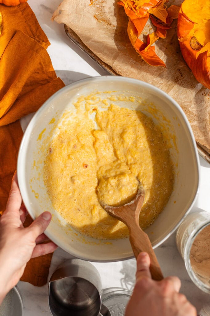 Stirring the PumpkinSourdough Base Hands stirring a vibrant yellow-orange mixture of pumpkin, sourdough starter, and other wet ingredients in a large white mixing bowl with a wooden spoon.