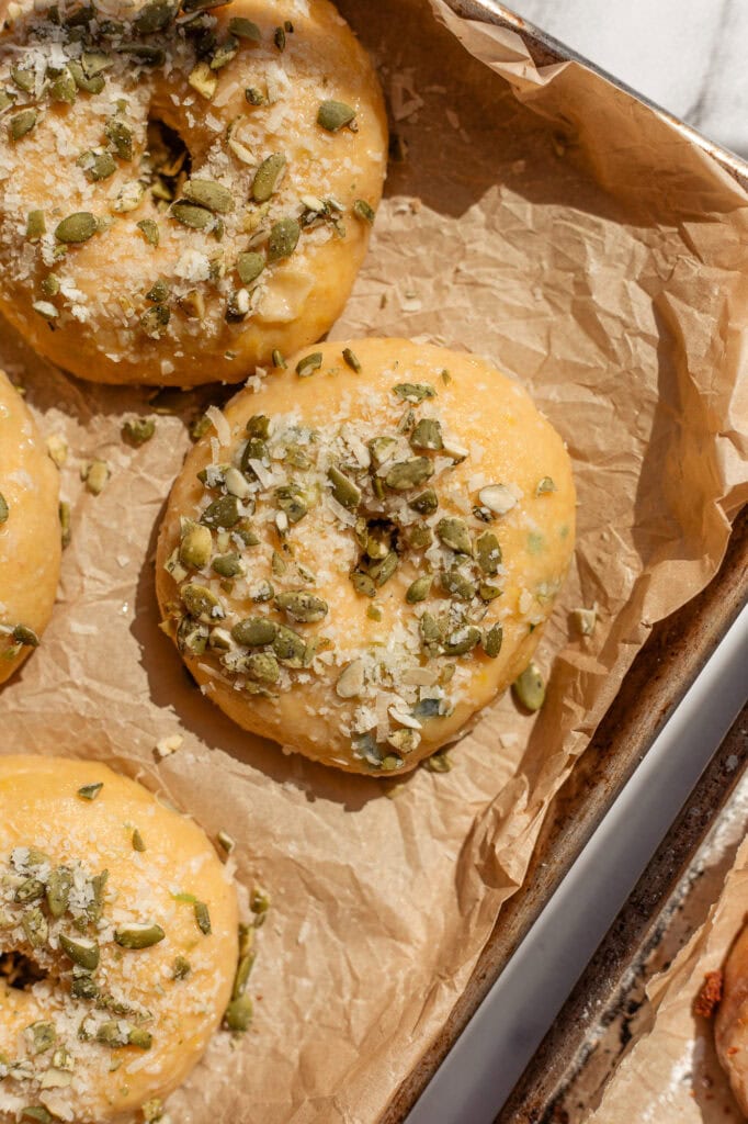 Topping the Bagels with Seeds and Parmesan A close-up of three savory pumpkin bagels resting on parchment paper, topped generously with pumpkin seeds, leeks and grated Parmesan.