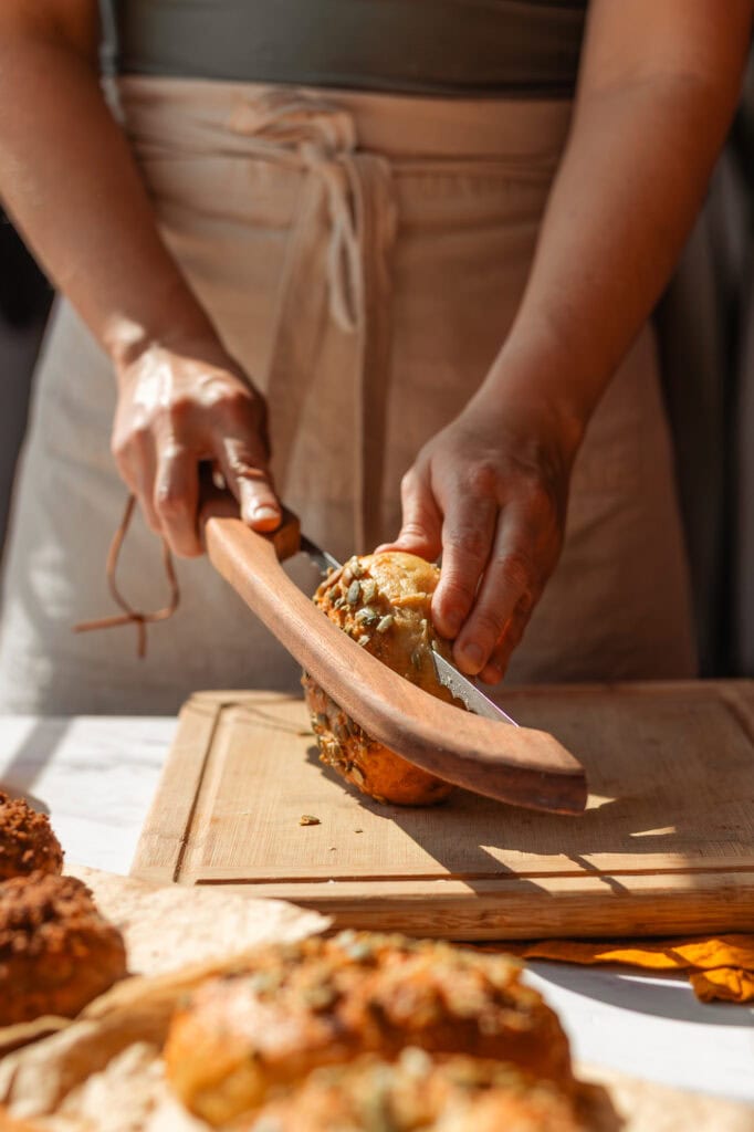 Slicing the Savory Roasted Pumpkin Bagel Hands using a wooden-handled bagel knife to slice a fully baked, golden-brown savory pumpkin bagel.