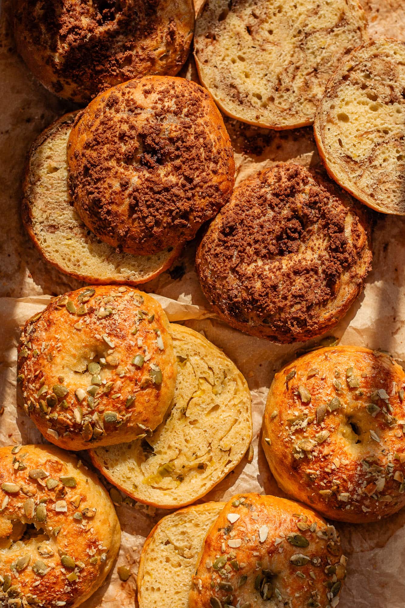 An overhead close-up of a batch of freshly baked sourdough roasted pumpkin bagels resting on parchment paper. The left side features four sweet cinnamon crumble bagels with a dark brown topping, and the right side features four savory bagels topped with vibrant green pumpkin seeds and parmesan.