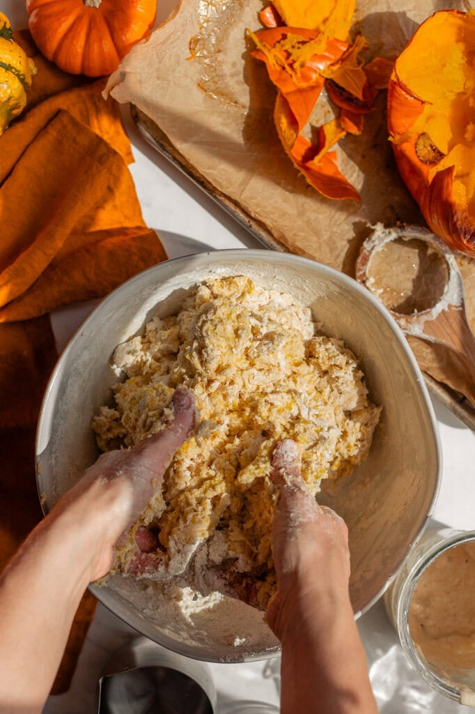 HandKneading the Pumpkin Bagel Dough Hands mixing and kneading a shaggy, yellow-orange pumpkin sourdough dough inside a large, silver metal mixing bowl.