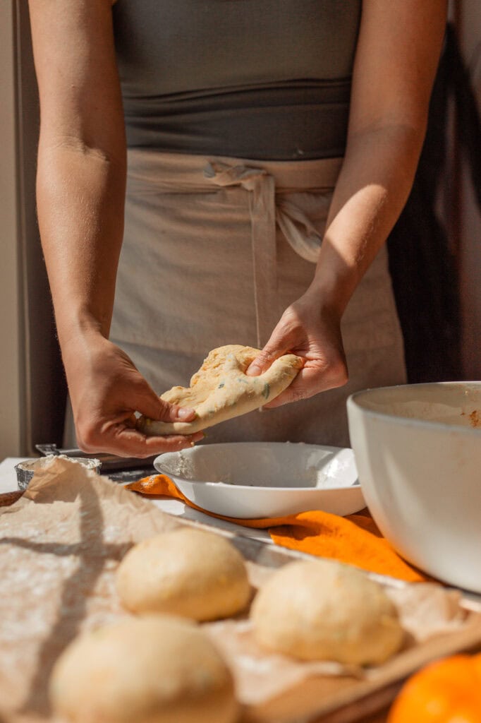 Rolling the Savory Bagel Dough into Balls Hands gently flattening and stretching a quarter portion of the pumpkin sourdough dough, preparing to roll it into a tight ball for the pre-shape step.