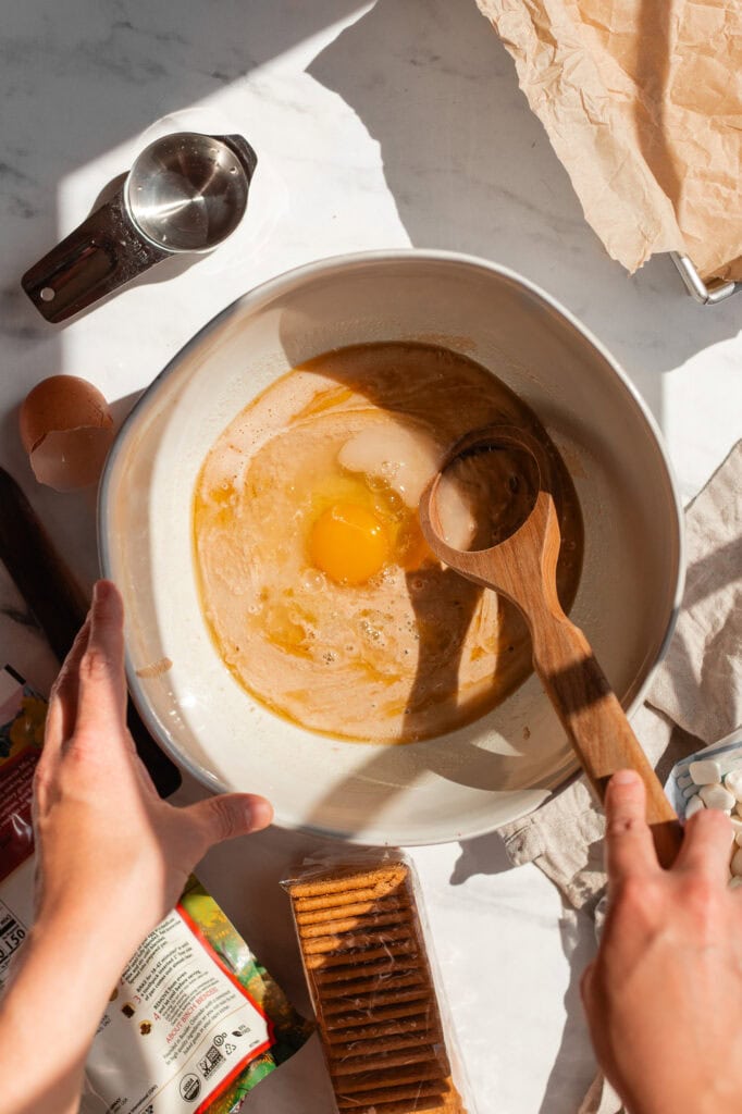 An egg is added to the browned butter and sourdough starter mixture. A wooden whisk is mixing the ingredients. 