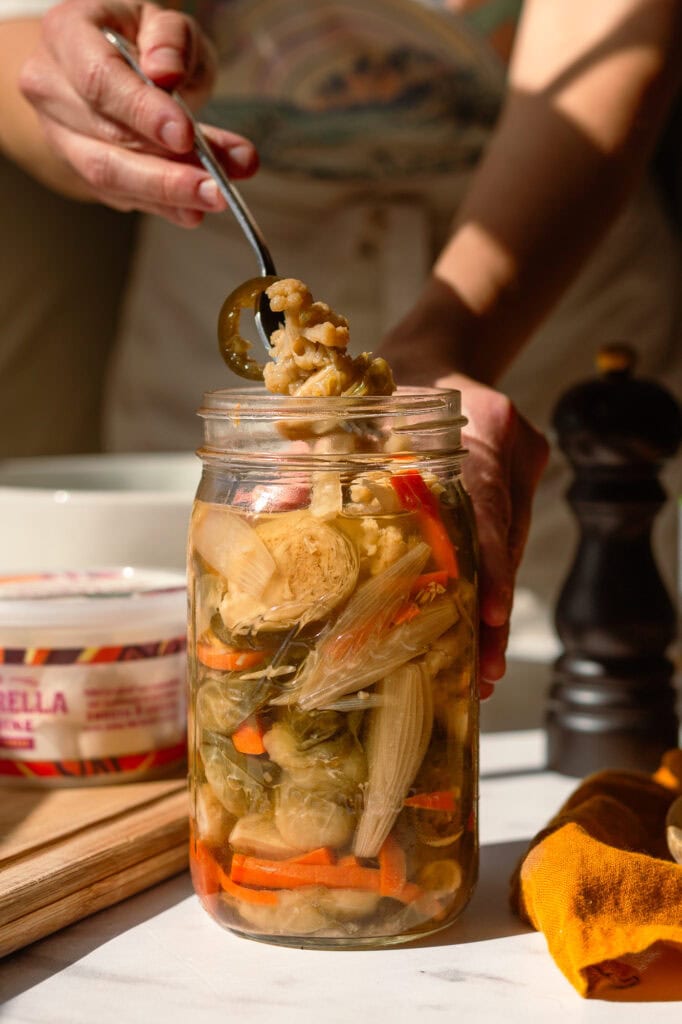 someone scooping fermented giardiniera out of a glass mason jar to chop as a main ingredient in Italian deli pasta salad.