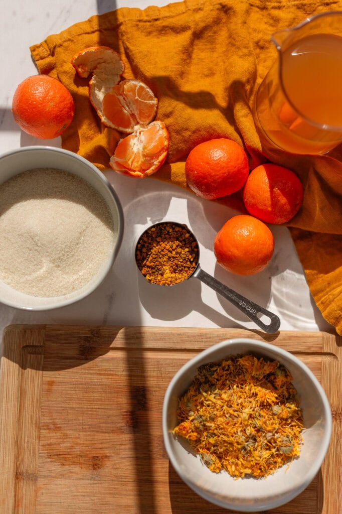 Shrub vinegar ingredients on a table, including 2 cups of vinegar, 1 cup of granulated sugar, 2 cups of mandarin oranges, and 1/2 cup of mixed herbs (turmeric and calendula)
