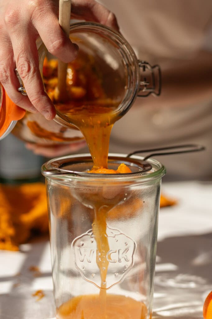 after mulling in the fridge for a few days, someone is holding a strainer over a clean jar and straining the fruit and herb pieces from the shrub vinegar.