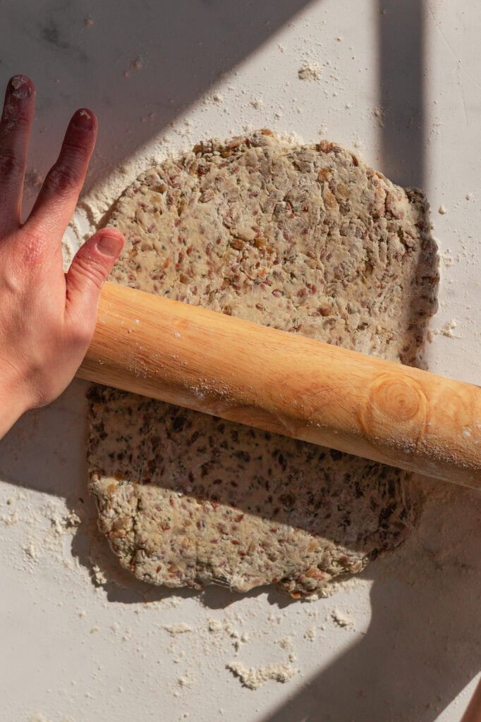 Hands using a rolling pin to flatten the sourdough starter and seed mixture.