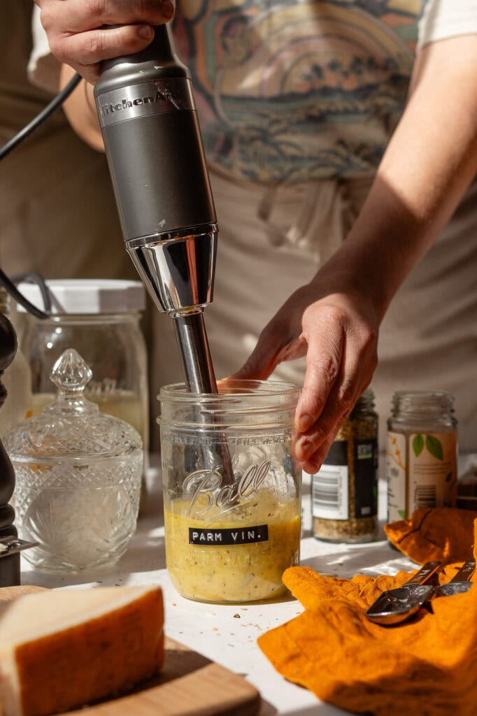 someone using a ktichen aid stick blender (aka an immersion blender) to blend the parmesan vinaigrette ingredients in a wide mouth glass mason jar. 