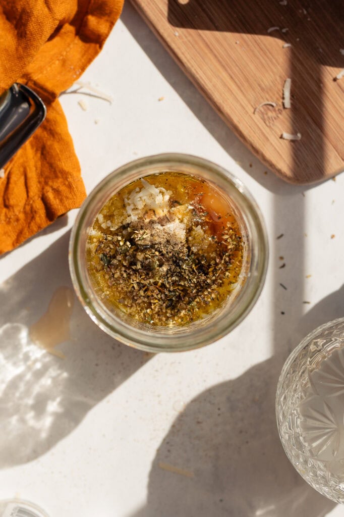 a wide mouth glass mason jar with olive oil, vinegar, pickle brine, spices, and parmesan cheese in it. It's about half full and ready to be blended into parmesan vinaigrette