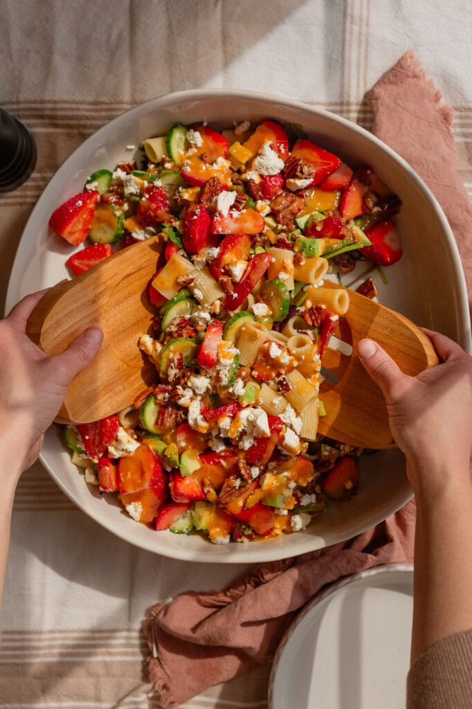 someone using wooden salad tongs to toss all the strawberry pasta salad ingredients together in a large ceramic bowl.