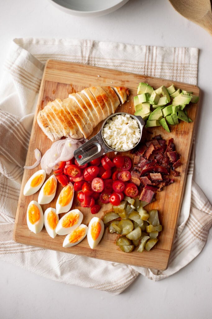 the main ingredients for Chicken Caesar Cobb Pasta Salad on a bamboo cutting board. There's sliced chicken, chopped bacon, crumbled feta, chopped fermented pickles, sliced cherry tomatoes, and thin sliced shallot. 