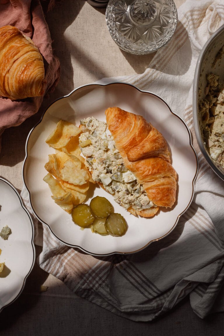 Croissant sandwich with a creamy chicken salad, potato chips, and pickles on a white plate, top-down view