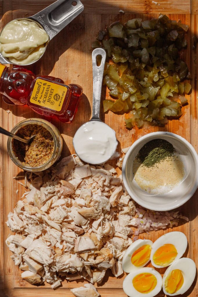 Chopped cooked chicken on a wooden cutting board with other dill pickle chicken salad ingredients: hard-boiled egg halves, yogurt, mayo, pickles and seasonings laid out for prep.