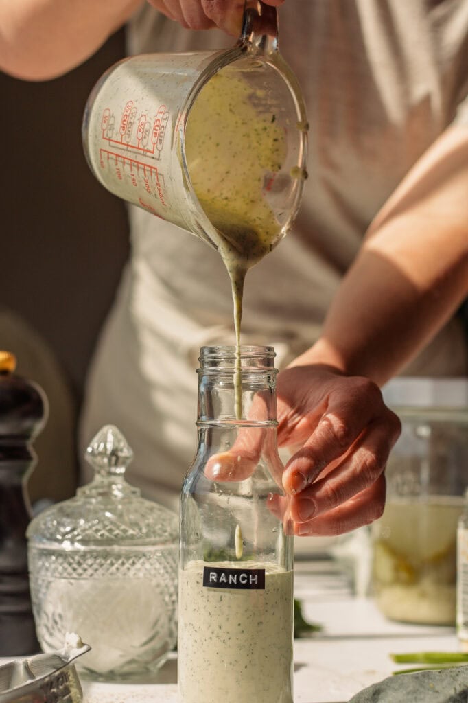 pouring blended greek yogurt ranch dressing from the glass measuring cup into a glass salad dressing bottle labeled "ranch"