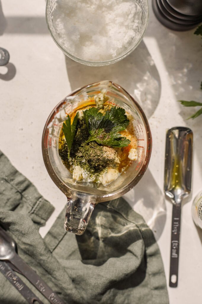 the ingredients to make greek yogurt ranch dressing in an 8- ounce liquid measuring cup before blending. There are fresh parsley leaves and cracked black pepper on top of the yogurt and olive oil.