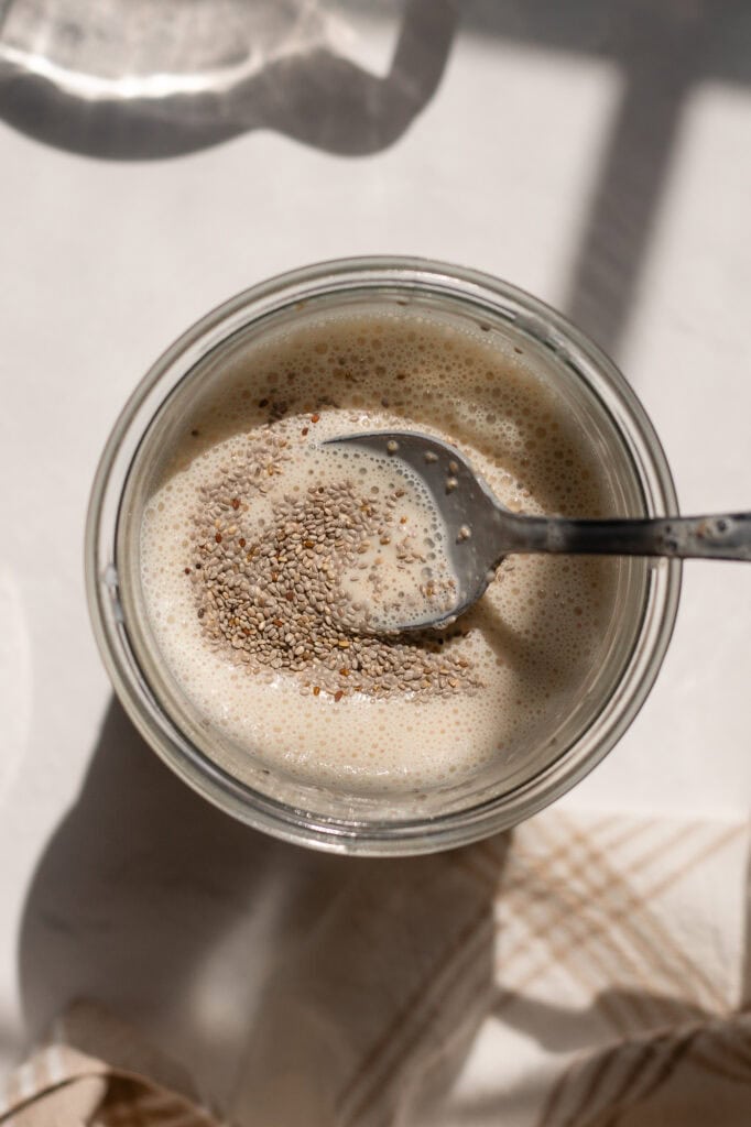 someone using a stainless steel spoon to stir white and grey chia seeds into a liquid mix or yogurt, milk, peanut butter, maple syrup, and vanilla. This is the base for the peanut butter yogurt chia pudding.