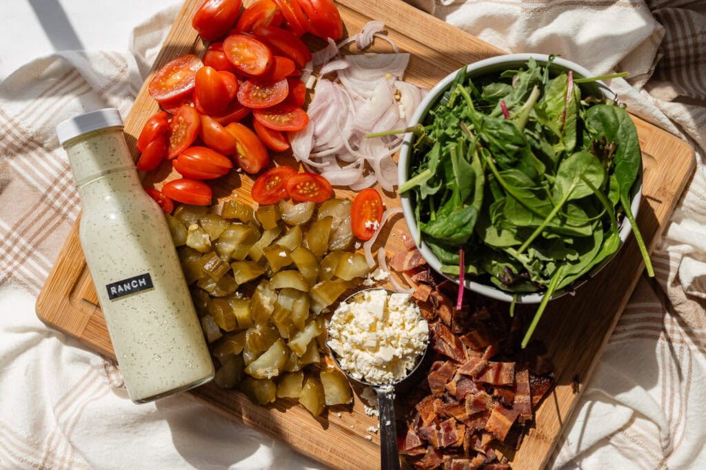 measured out ingredients for pickle bacon ranch pasta salad on a wooden cutting board. from left to right there is a bottle of ranch, chopped cherry tomatoes, chopped pickles, sliced shallot, crumbled feta, leafy greens, and roughly chopped bacon. 