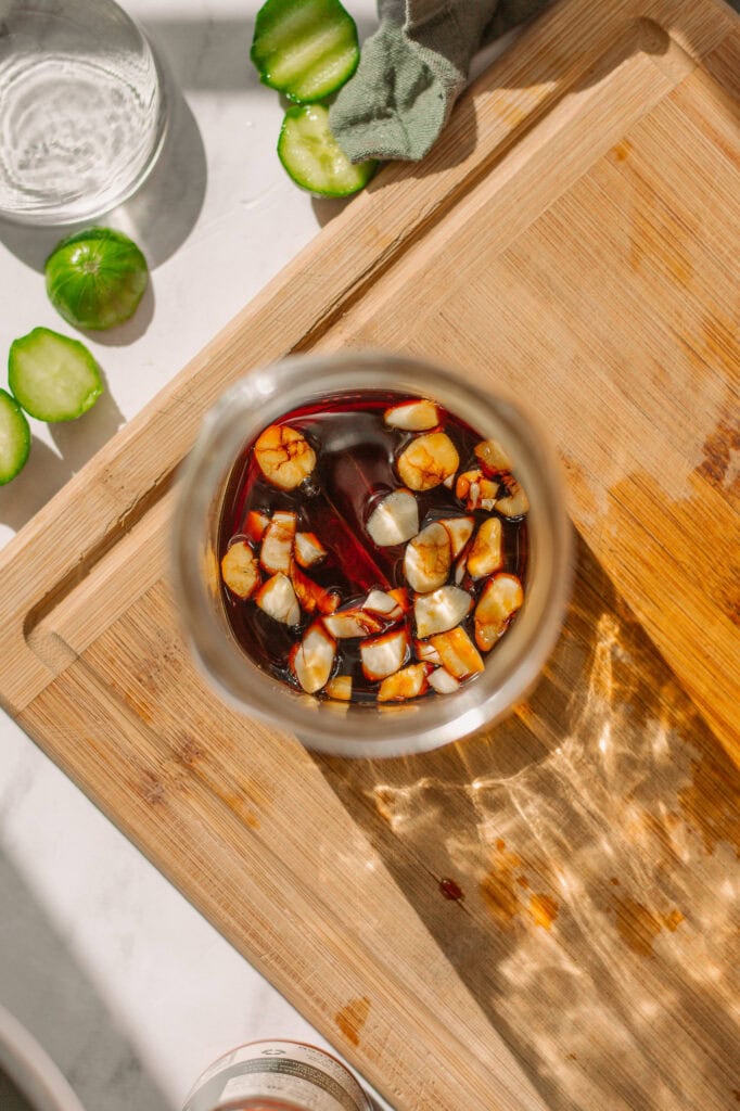 Worcestershire sauce and crushed garlic cloves in the bottom of a mason jar, the first ingredients added to the jar to make Blooody Mary Pickles.