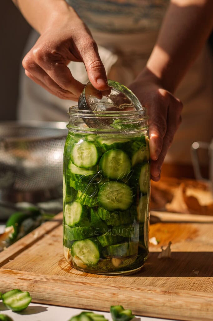 someone using their washed hands to place a wide-mouth glass fermentation weight into a wide mouth, 32 ounce, glass mason jar filled with chopped cucumbers, roasted garlic, roasted jalapenos and dill.