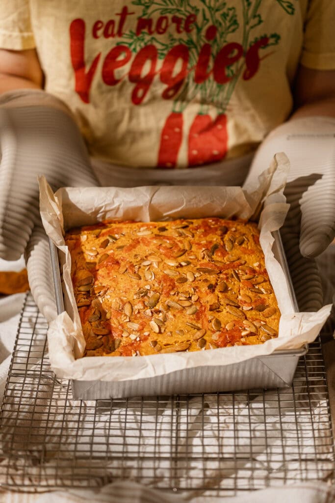 someone with oven mats on placing the pumpkin cornbread, still in the pan, on a cooling rack to cool in the pan for 10 minutes.