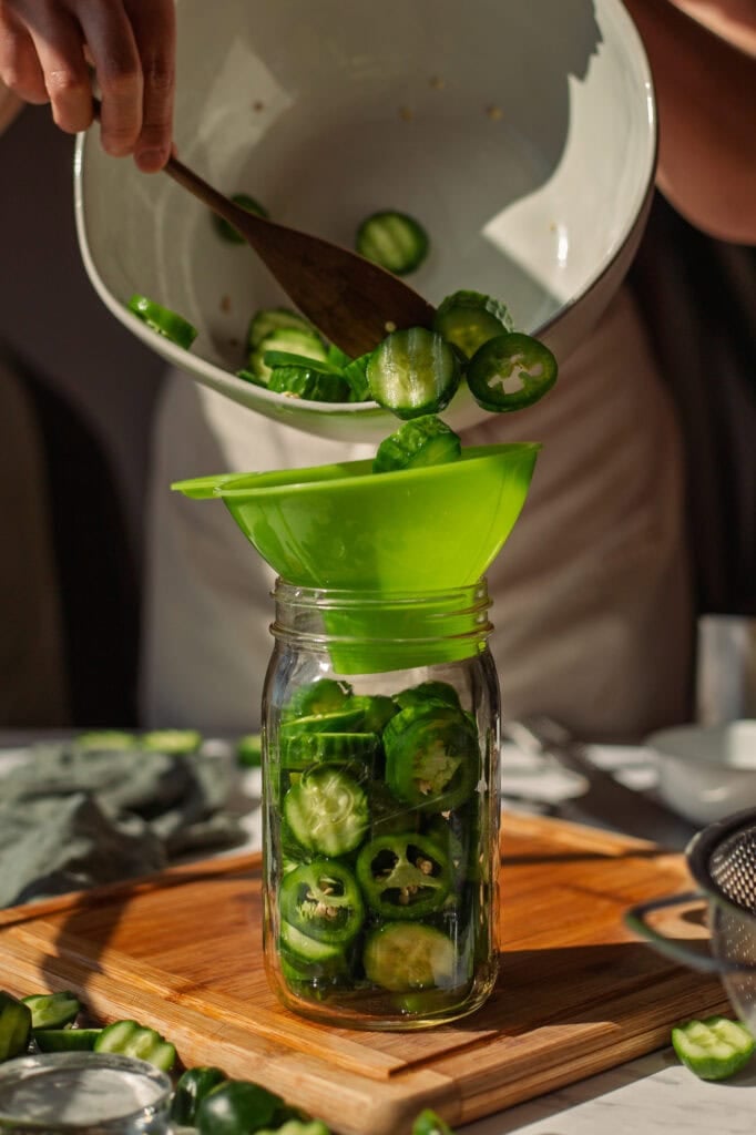 A funnel is used to pour cucumbers and jalapeño slices into a glass mason jar.