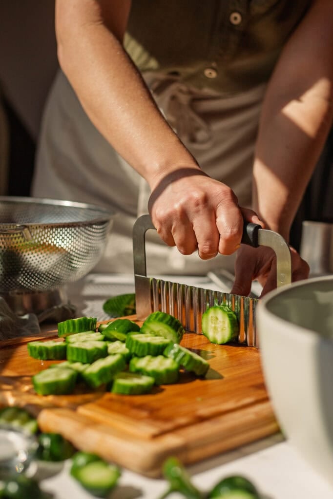 hands use a slicer to cut cucumbers into slices on a wooden cutting board.